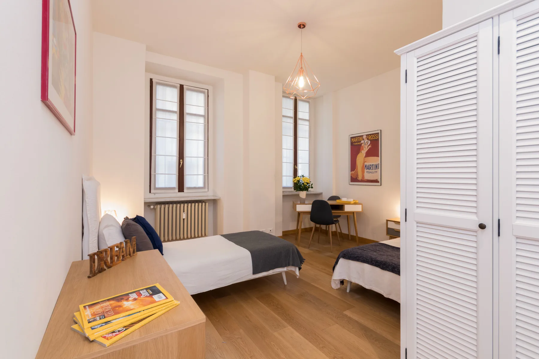 Un dormitorio luminoso con dos camas, ventanas altas y un escritorio de madera.