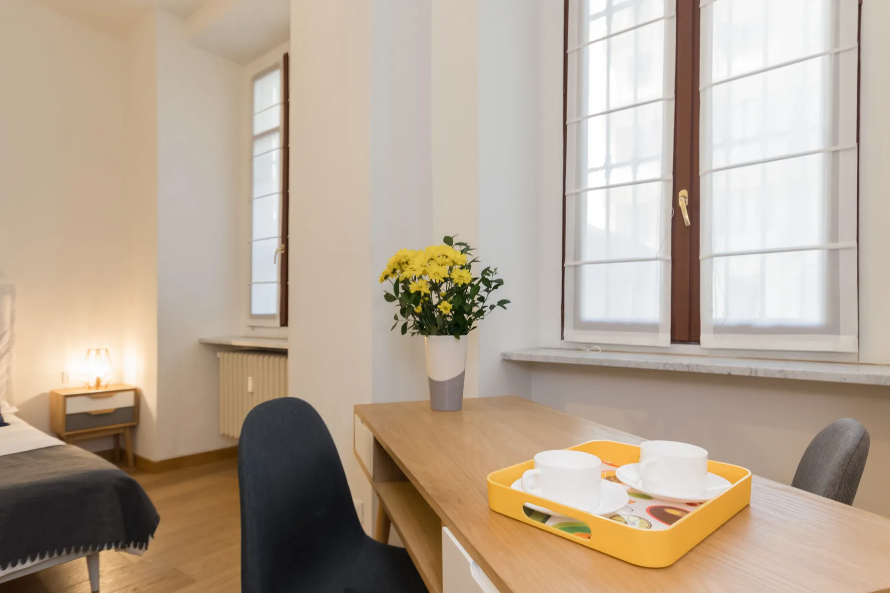Un dormitorio luminoso con una mesa de madera, una bandeja con tazas y flores junto a la ventana.