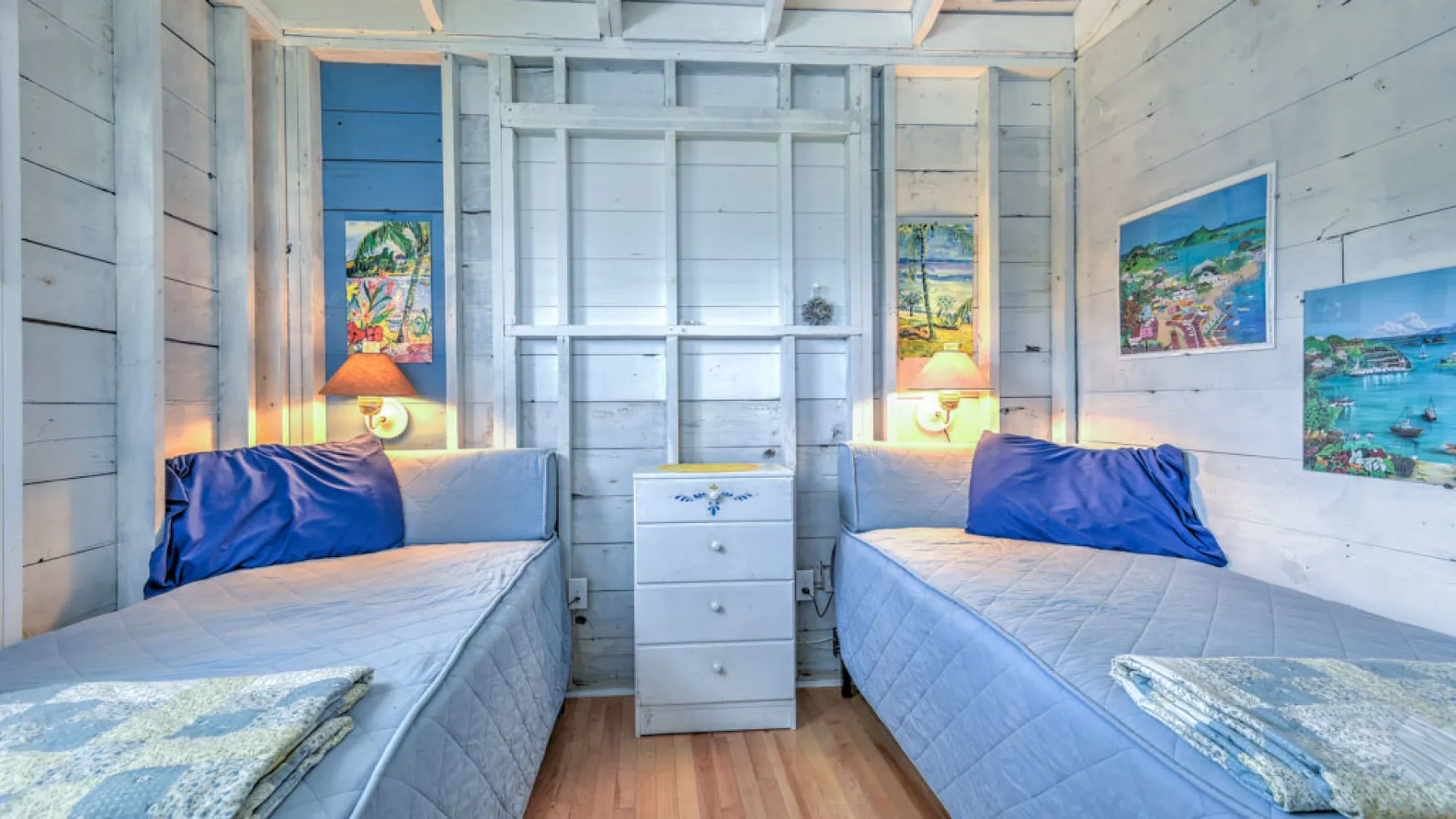 A bright bedroom with two beds, a central dresser, and colorful wall art.