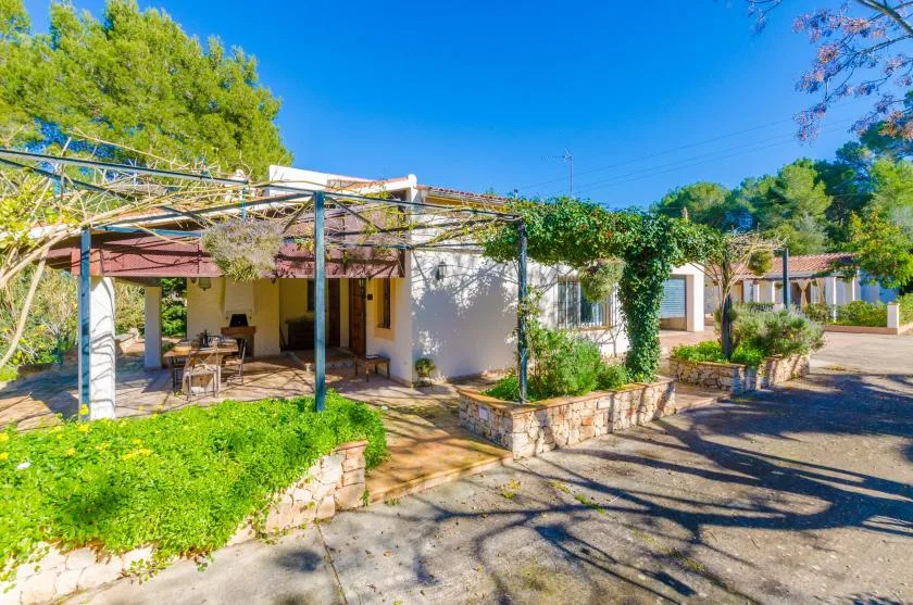 Una casa baja con un patio sombreado está rodeada de vegetación junto a una entrada pavimentada.