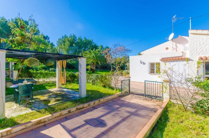Un patio ajardinado junto a una casa blanca, con una zona de estar exterior a la sombra.