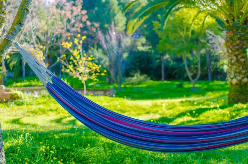 Una hamaca a rayas cuelga sobre un jardín verde bajo árboles frondosos.