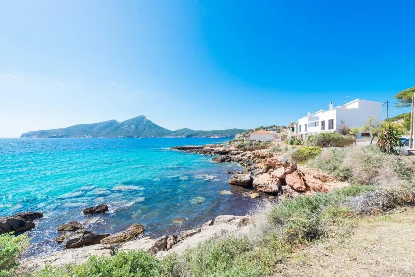 Una costa rocosa bordea aguas azules y claras, con edificios blancos junto al mar.