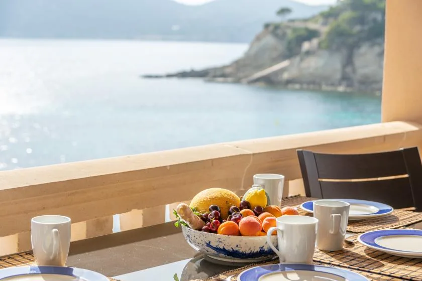 Una mesa de desayuno con fruta y tazas mira al agua y la costa rocosa.