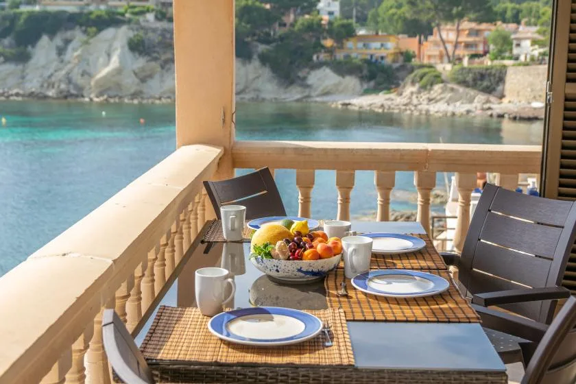 Una mesa de comedor en el balcón da a aguas turquesas y una costa rocosa.