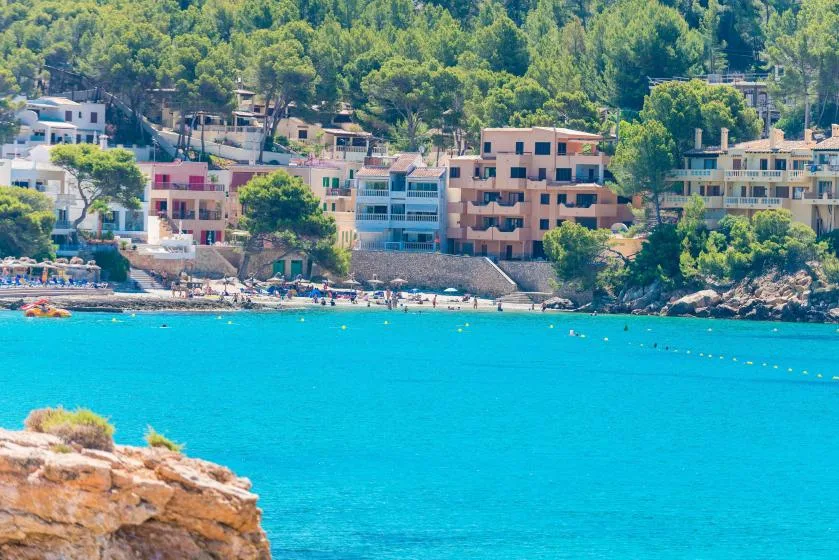 Una bahía turquesa bordea una playa y edificios junto al agua, bajo una ladera verde.