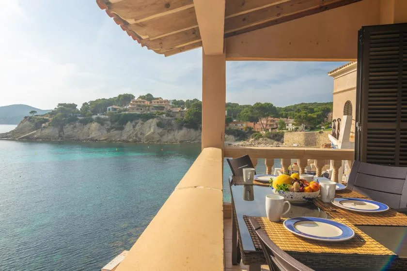 Un balcón cubierto con una mesa de comedor da al agua y a una costa rocosa.