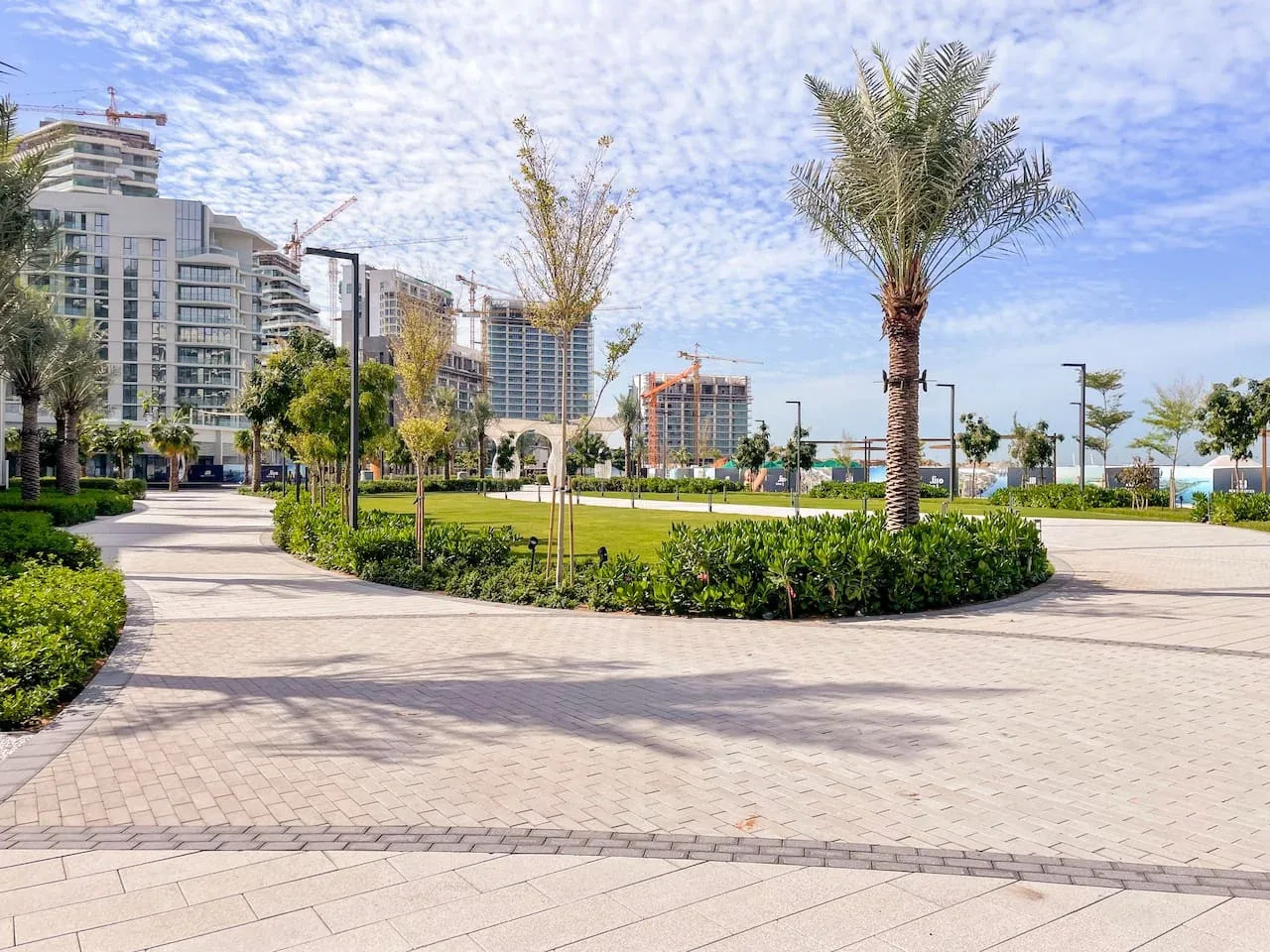 Parque urbano ajardinado con senderos pavimentados, árboles jóvenes y edificios modernos.