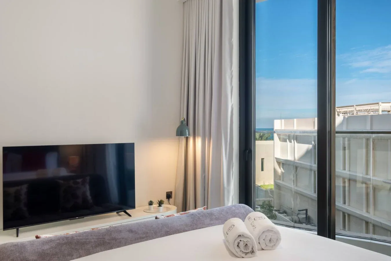 Un dormitorio con un televisor junto a ventanas altas con vistas a edificios cercanos.