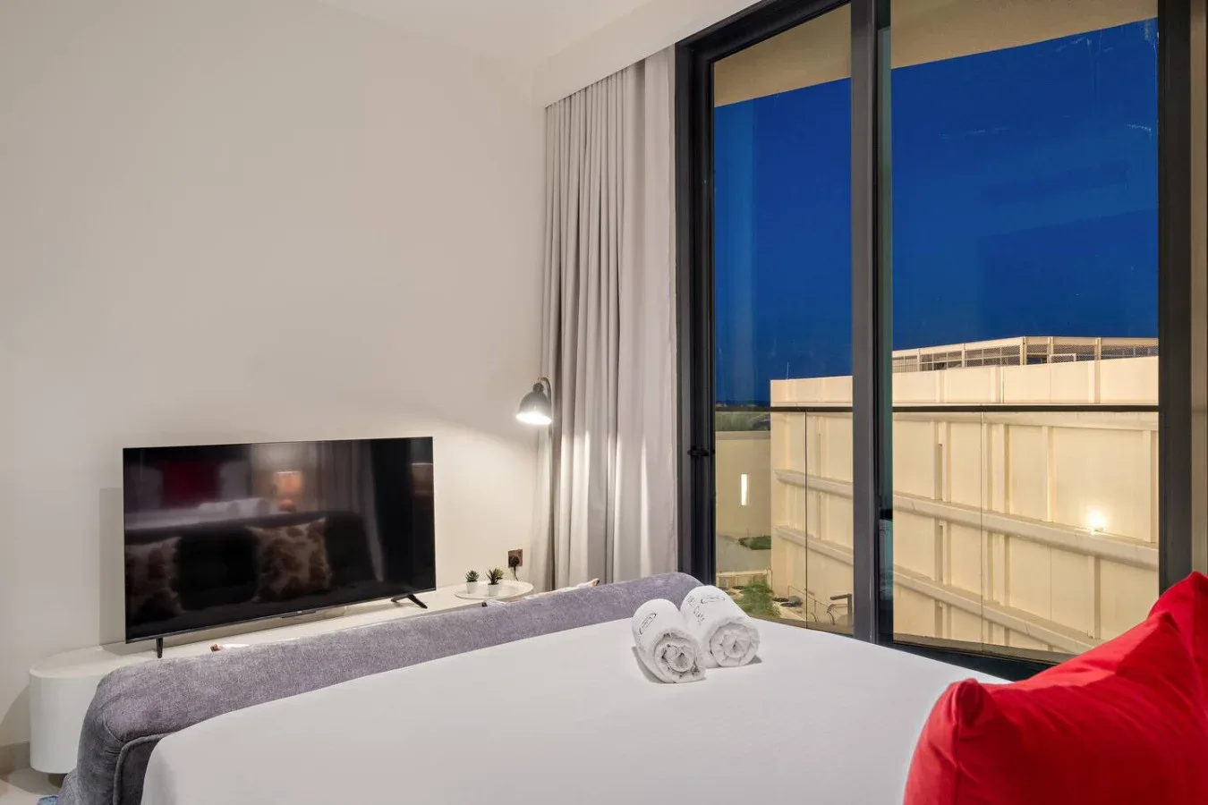 Un dormitorio con una cama junto a ventanales que dan a un edificio iluminado.