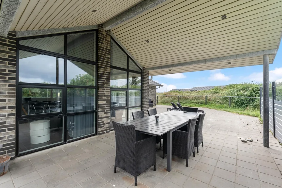 Una zona cubierta de comedor al aire libre con mesa y asientos junto a grandes ventanas.