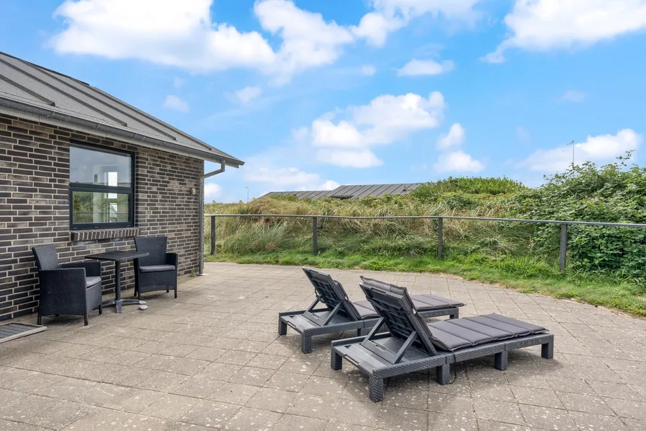 Un patio pavimentado junto a un edificio de ladrillo, con asientos y vegetación al fondo.