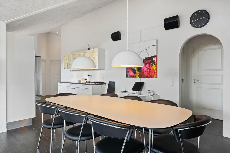 Un comedor luminoso tiene una mesa larga, lámparas colgantes y una cocina al fondo.