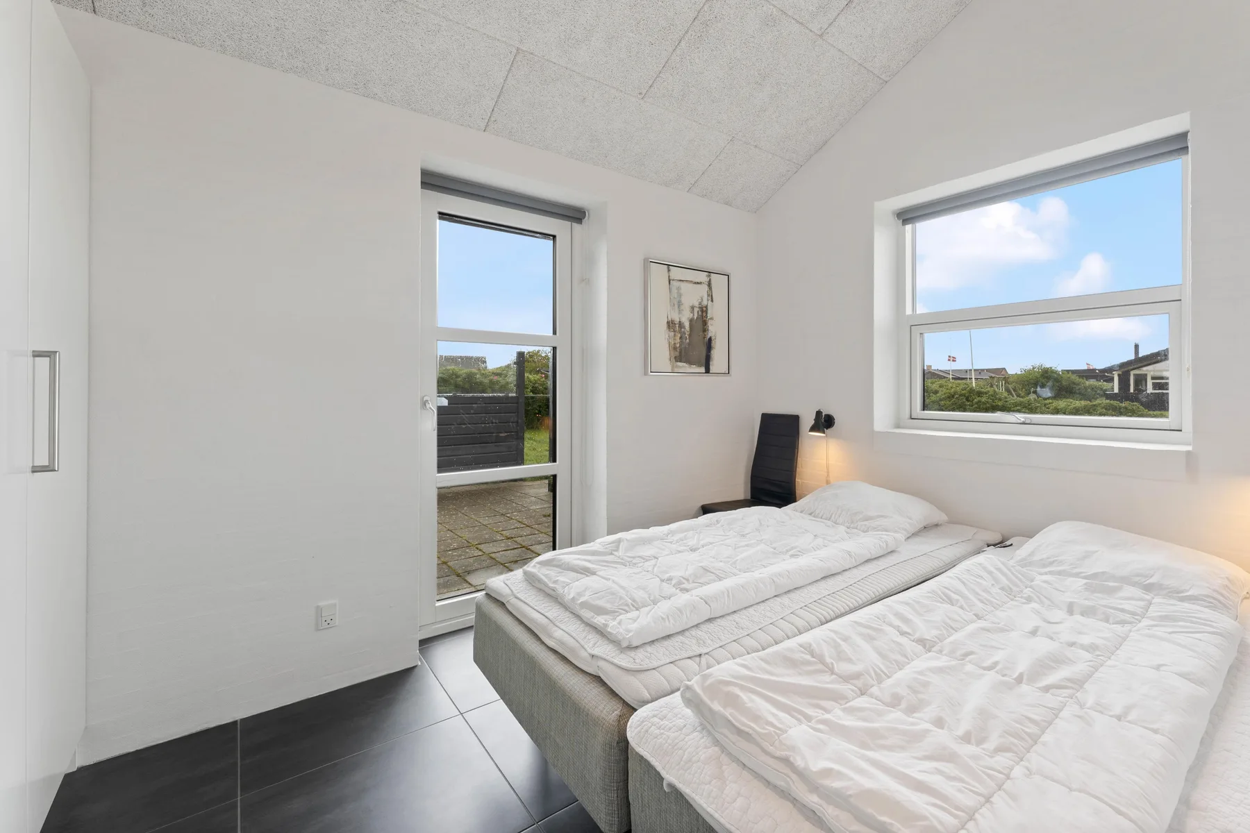 Un dormitorio luminoso con camas, ventanas amplias y una pequeña lámpara de noche.