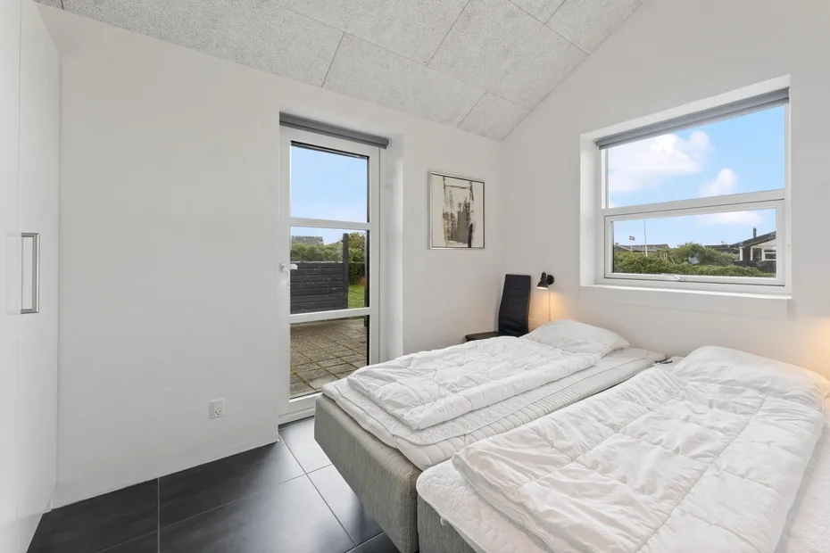 Un dormitorio luminoso con dos camas, ropa de cama blanca y ventanas grandes.