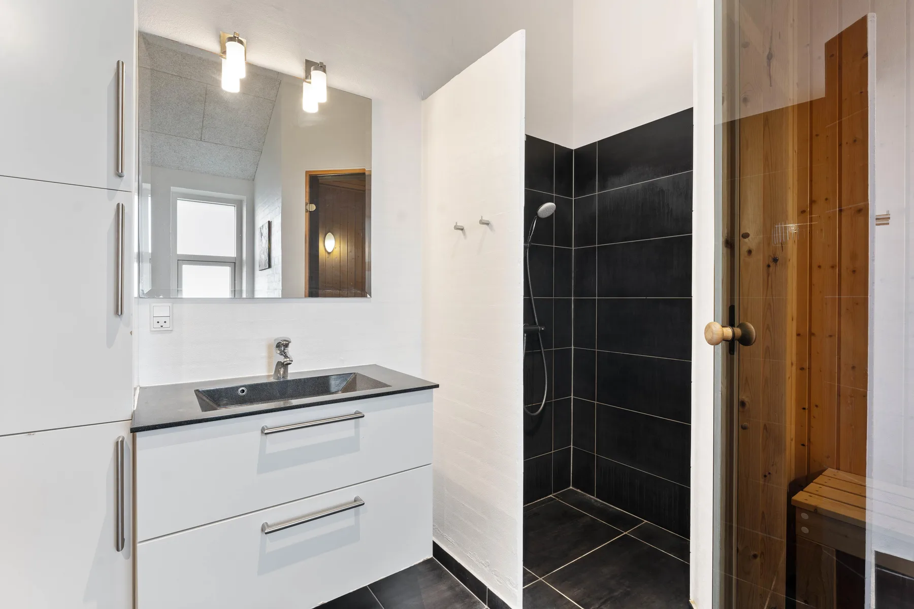 Un tocador de baño y un espejo están junto a una ducha con azulejos oscuros y una puerta de madera.