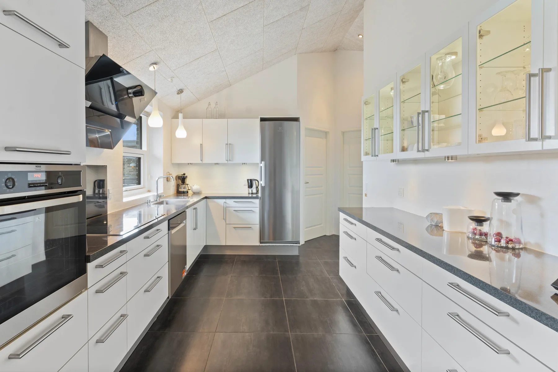 Una cocina moderna y luminosa con gabinetes blancos, electrodomésticos de acero inoxidable y suelo de baldosas oscuras.