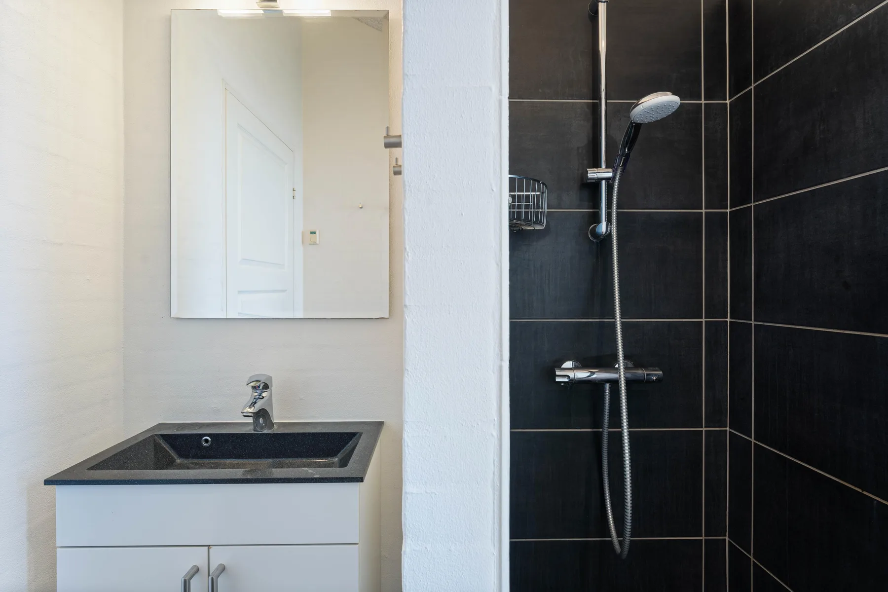Un baño con lavabo y espejo junto a una ducha de azulejos oscuros.