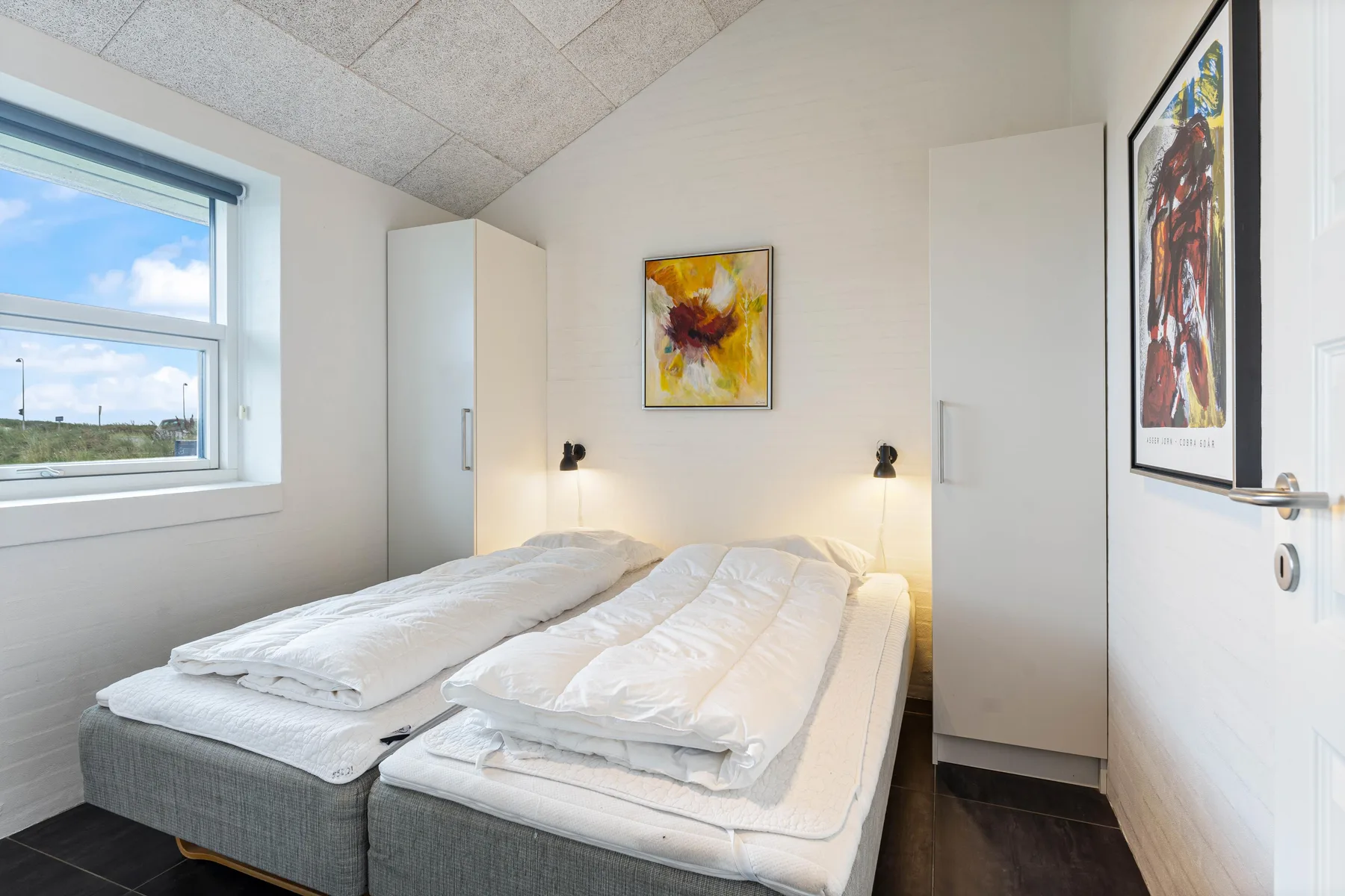 Un dormitorio luminoso con dos camas, ropa de cama blanca y cuadros enmarcados.