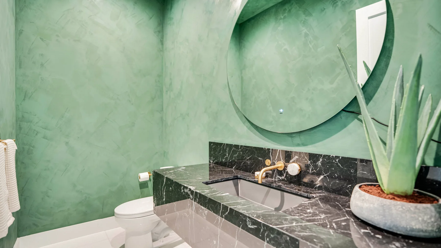 Un baño verde con lavabo, inodoro y un gran espejo redondo.