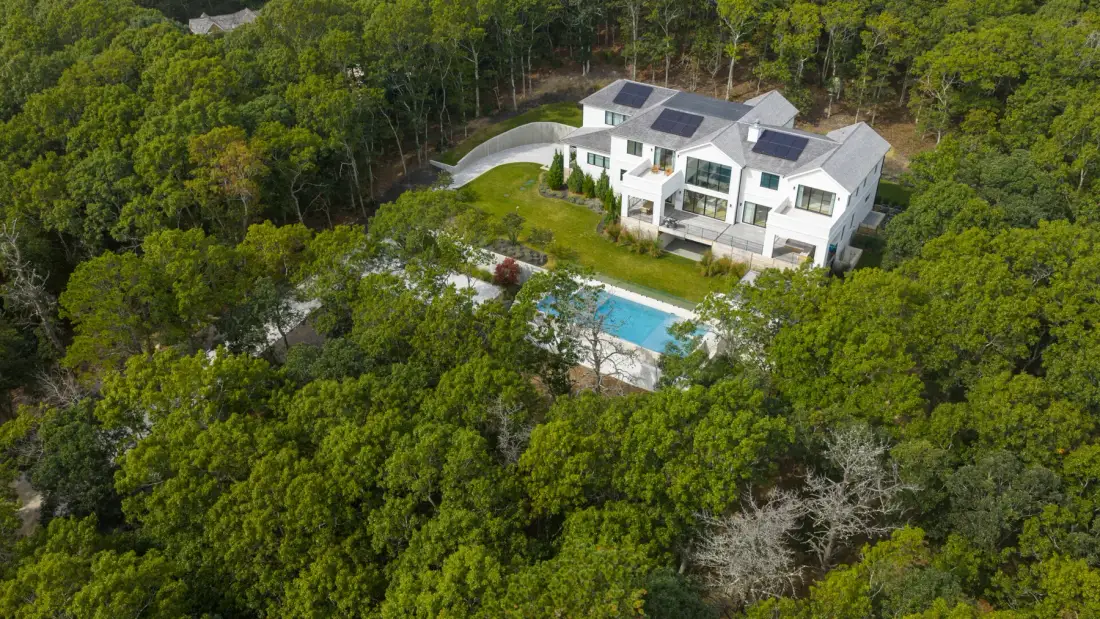 Una casa blanca con paneles solares y una piscina está rodeada de árboles densos.