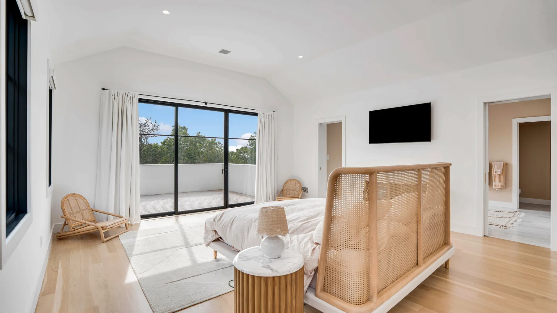 Un dormitorio luminoso con una cama, asientos de mimbre y puertas corredizas de vidrio hacia un balcón.