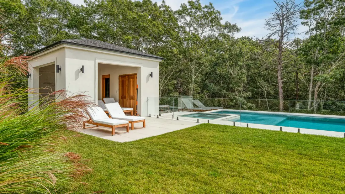 A small poolside building and lounge seating overlook a swimming pool amid trees.