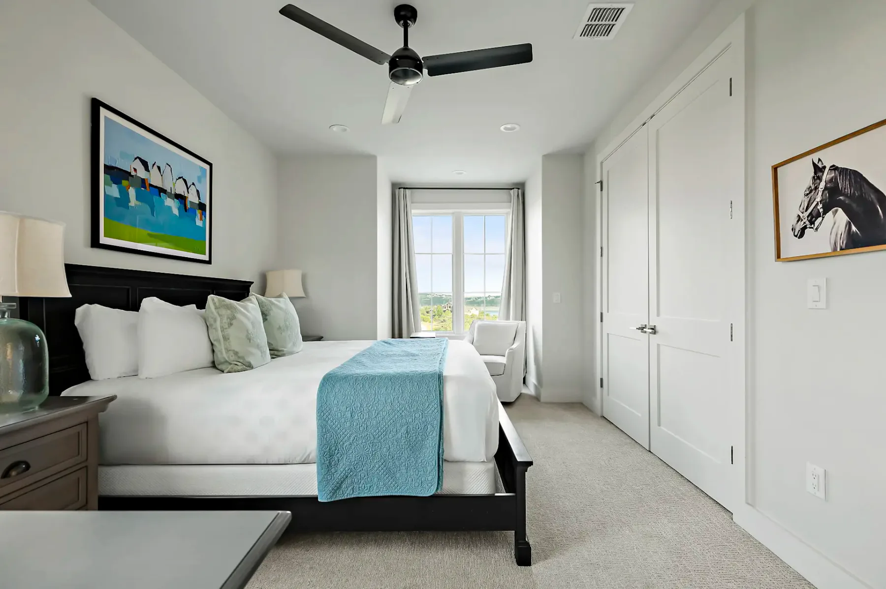 Modern bedroom with a large bed, blue runner, landscape painting, ceiling fan, and window overlooking greenery. Walls are light-colored.