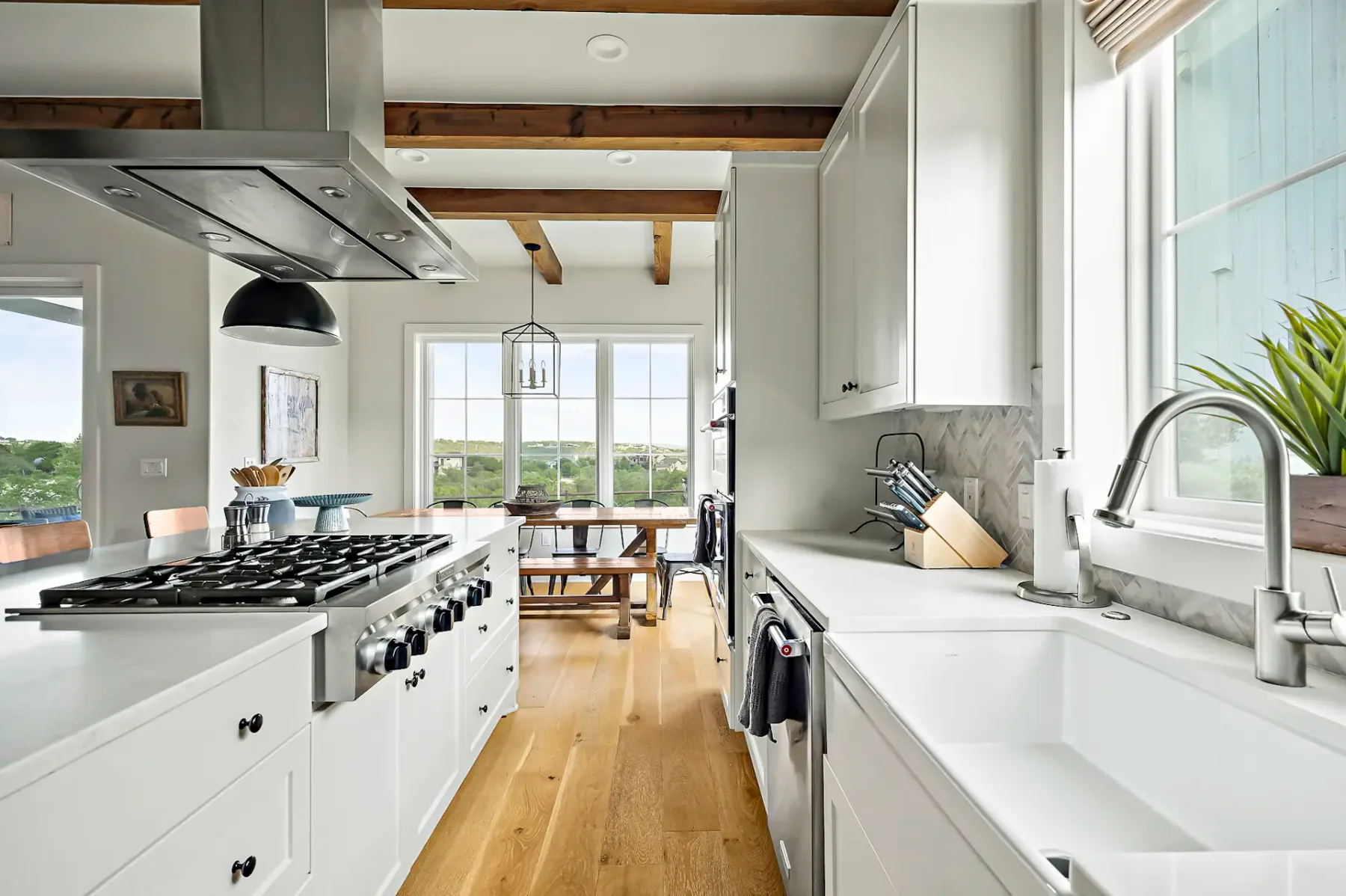 Modern kitchen with white cabinets, stainless steel appliances, wooden beams, and a farmhouse sink. Dining area visible through large windows.