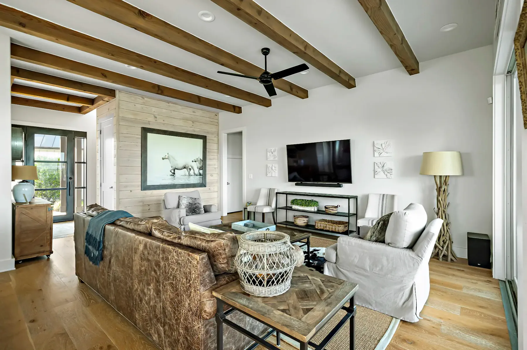 Stylish living room with wood beams, leather sofa, armchair, mounted TV, rustic decor, artwork of a horse, and large windows for natural light.