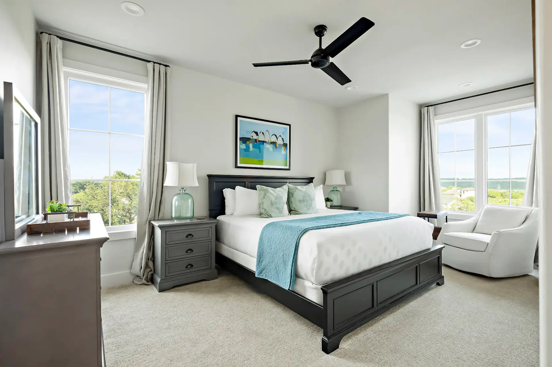 Modern bedroom with a large bed, teal accents, ceiling fan, two windows, bedside tables, and a landscape painting above the headboard.