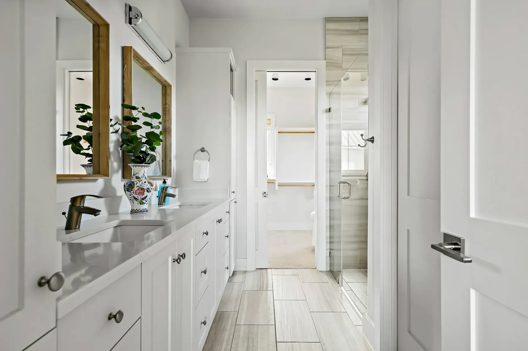 Bright, modern bathroom with dual sinks, large mirrors, a leafy plant, and a glass-enclosed shower. White cabinetry and neutral tiles featured.