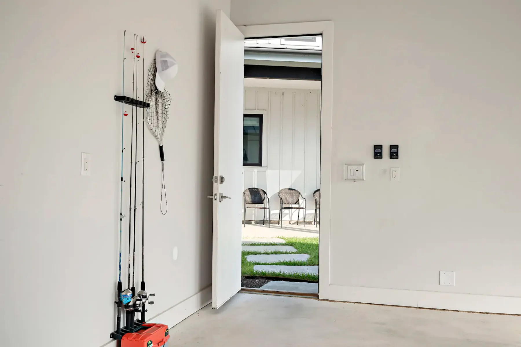 Open door in a garage leads to a patio with chairs and a pathway. Fishing rods and net are mounted on the wall.