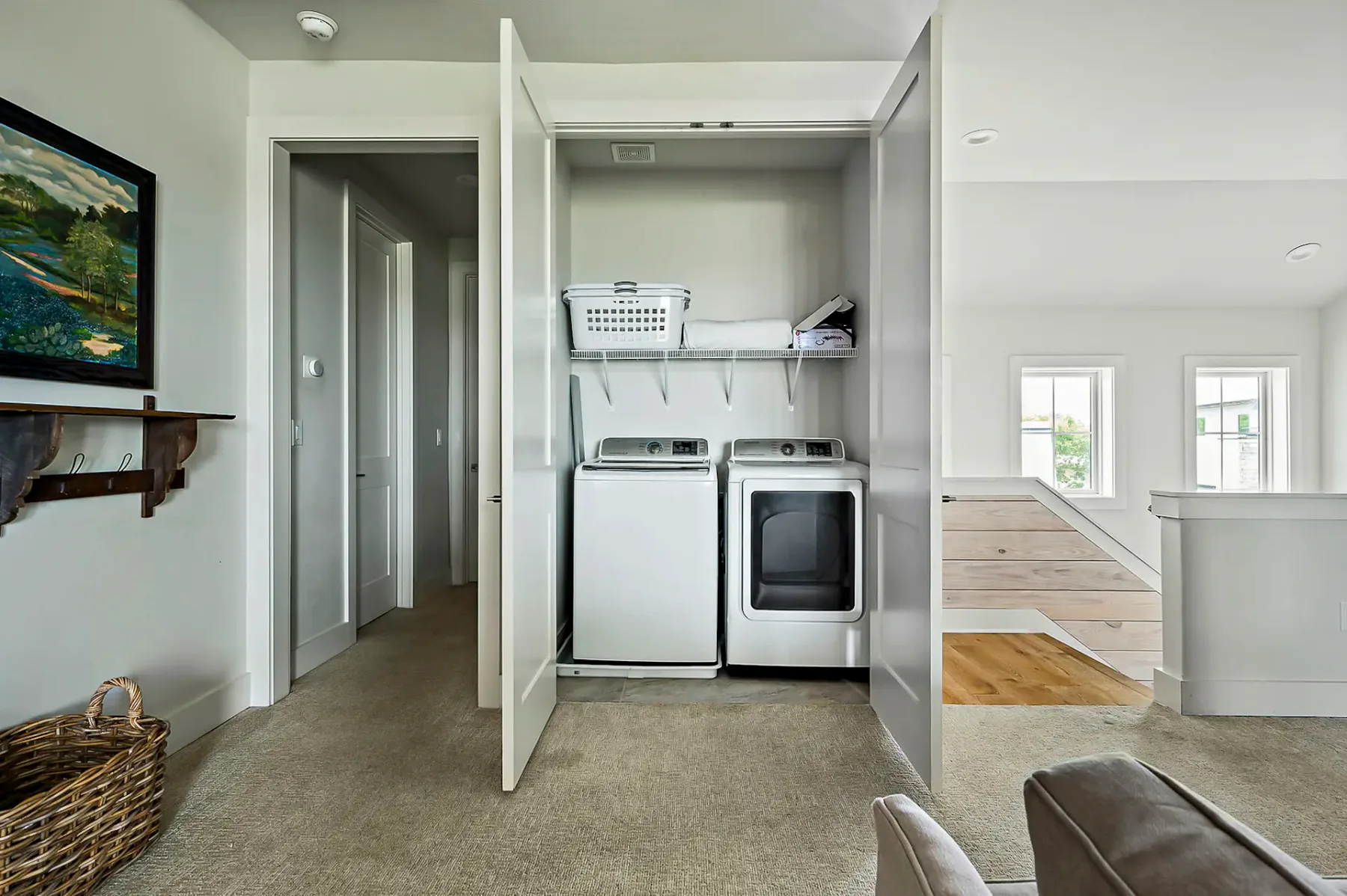 Laundry area with open double doors showing a washing machine, dryer, and a shelf holding a laundry basket. Staircase and framed art nearby.