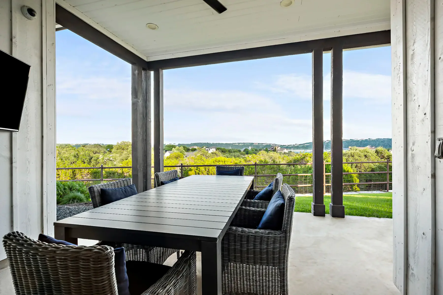 Covered patio with modern wicker furniture, long table, and scenic view of greenery and distant hills under a partly cloudy sky.