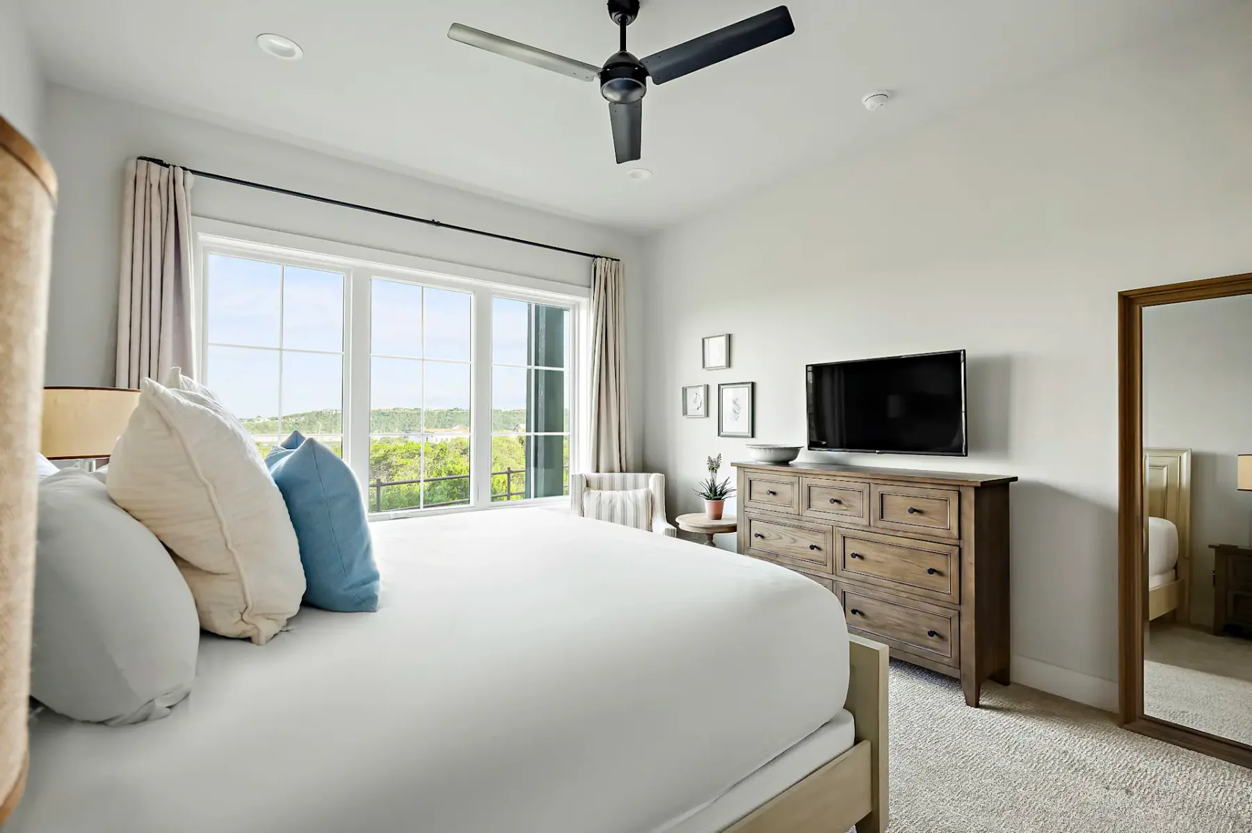 Bright bedroom with a neatly made bed, wooden dresser, TV, and large window showcasing a scenic view. Ceiling fan and wall art add to decor.