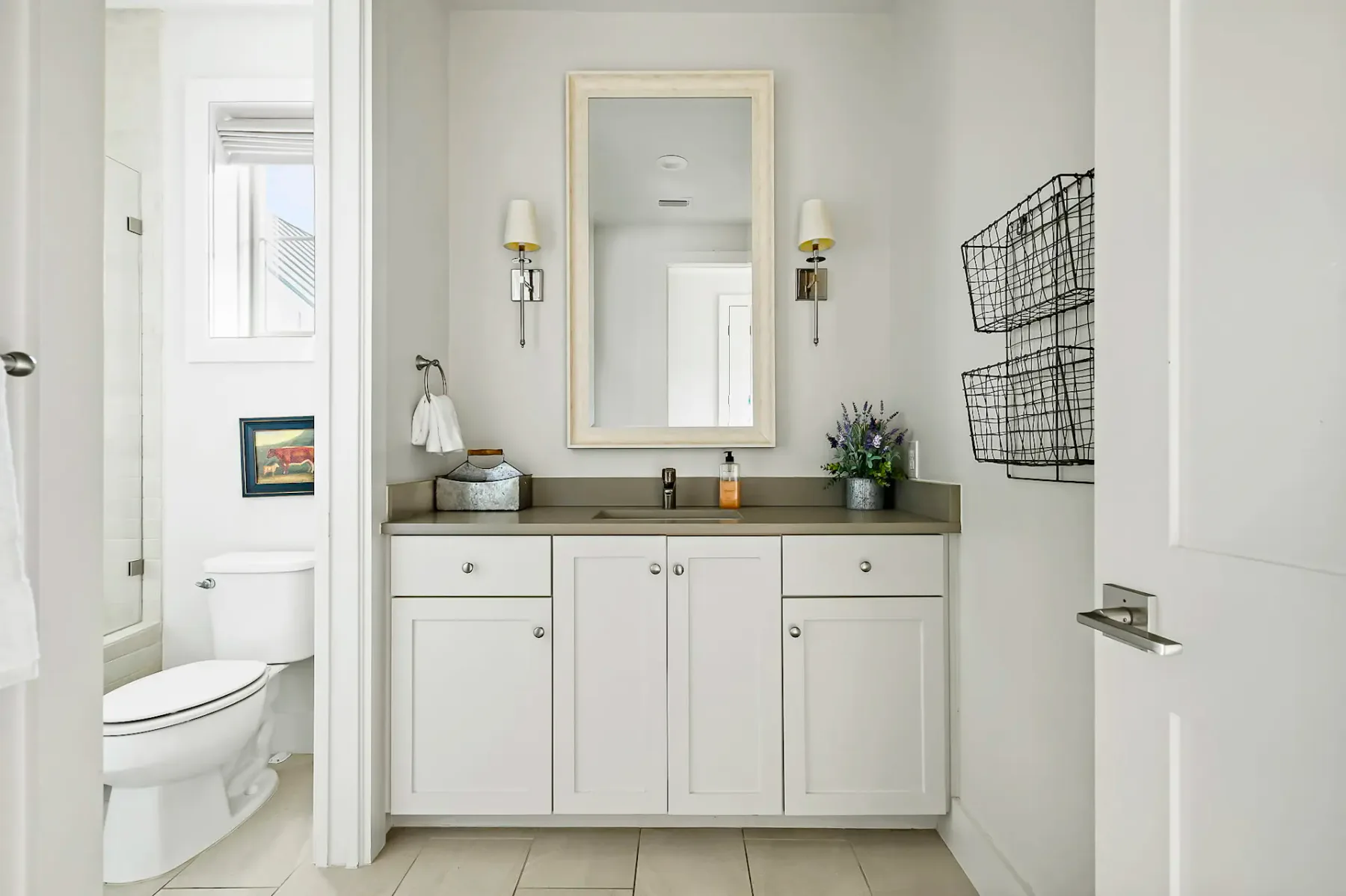A modern bathroom with white cabinets, a large mirror, wall sconces, and wire baskets. A toilet and small artwork are visible on the left.