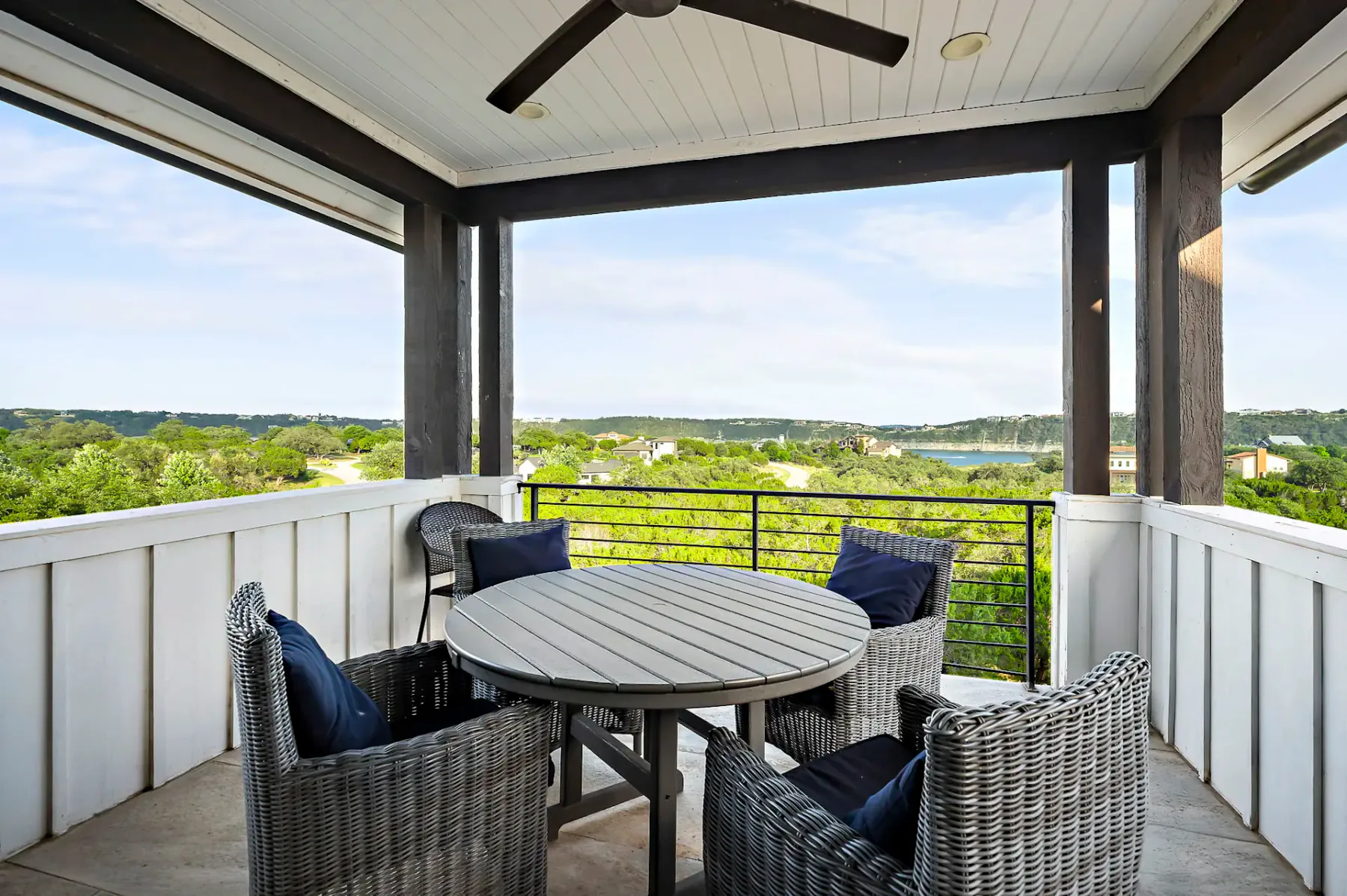 A covered balcony with a round table and four chairs overlooks a lush green landscape and distant water, under a ceiling fan.