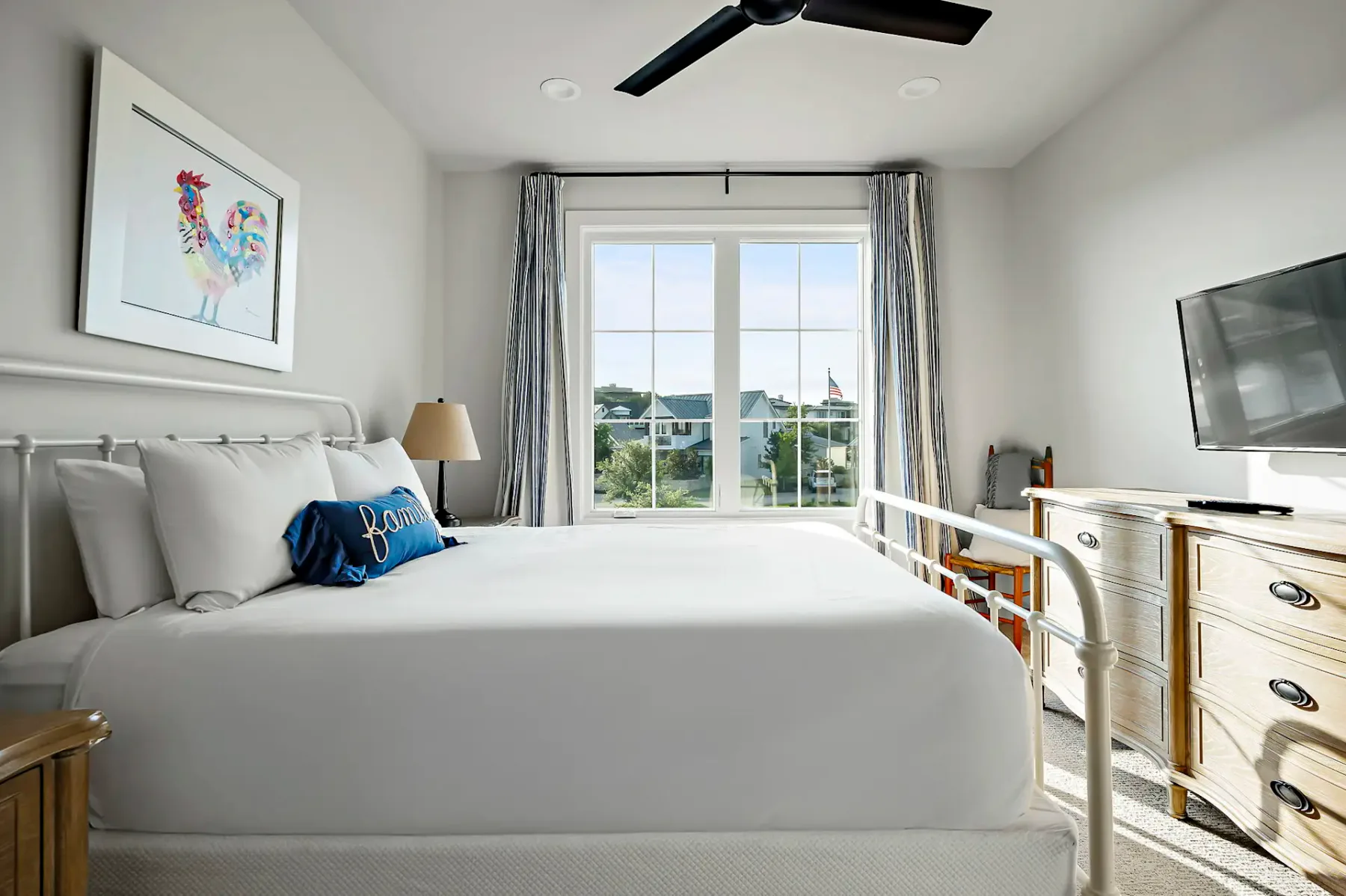 Bright bedroom with a white bed, rooster artwork on the wall, a TV on a wooden dresser, and a window with curtains overlooking a garden.