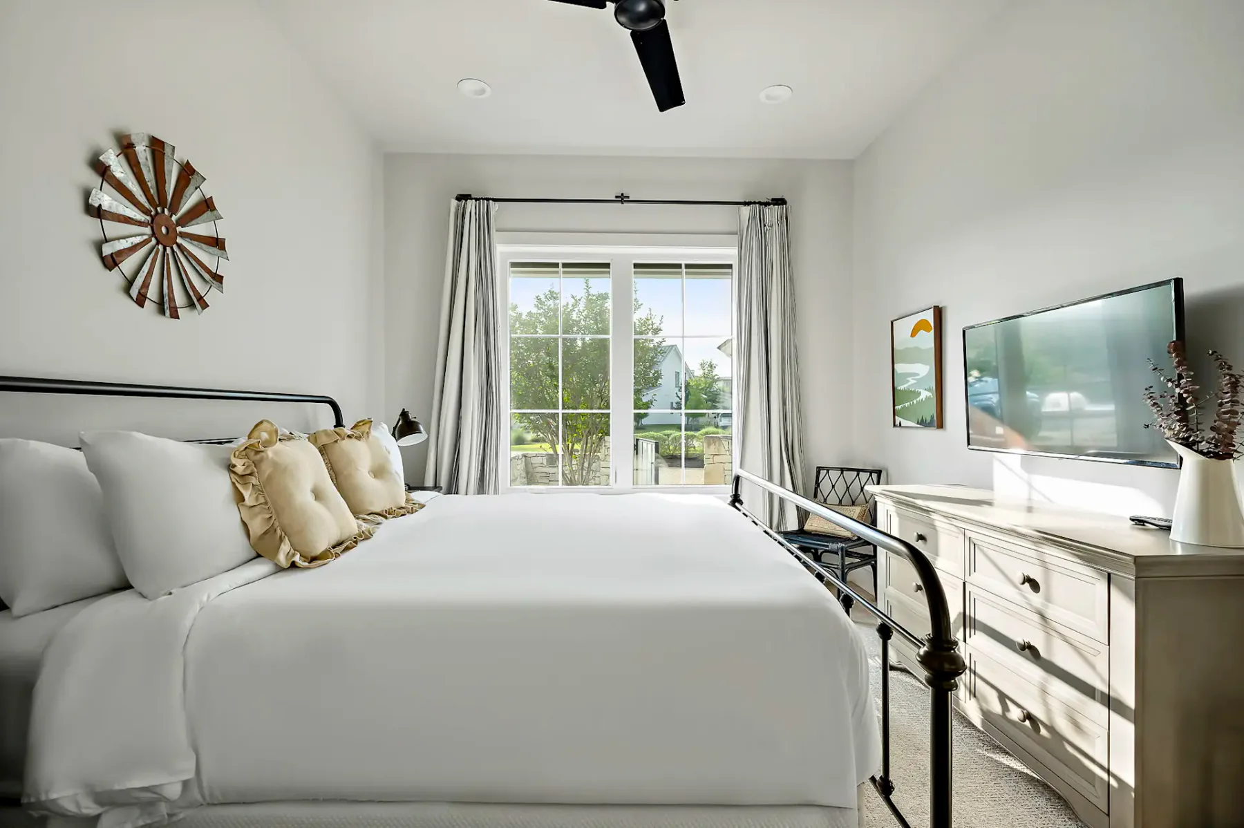 Bright bedroom with a metal bed, soft pillows, and a TV on a dresser. Large window with curtains offers a view of greenery outside.