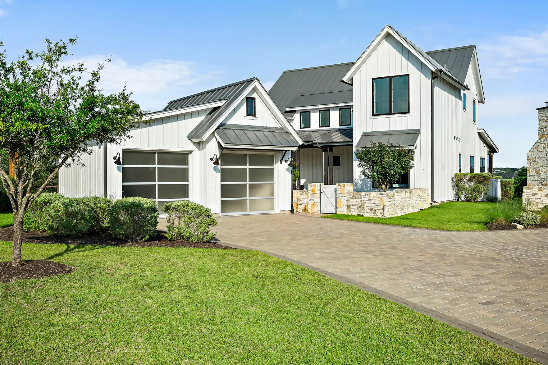 Modern white house with a gray roof, large windows, and a stone accent wall. Features a manicured lawn and a paved driveway.