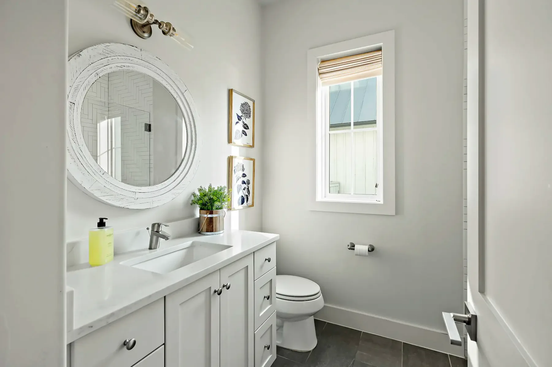 Modern bathroom with a white vanity, round mirror, potted plant, wall art, and a window with blinds. Minimalist decor and neutral colors.