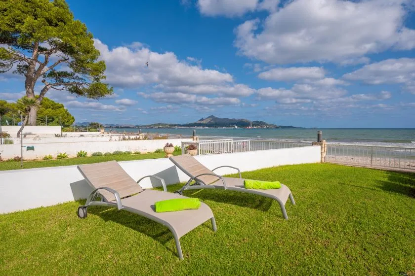 Outdoor loungers sit on a grassy lawn beside a waterfront, with hills across the water.