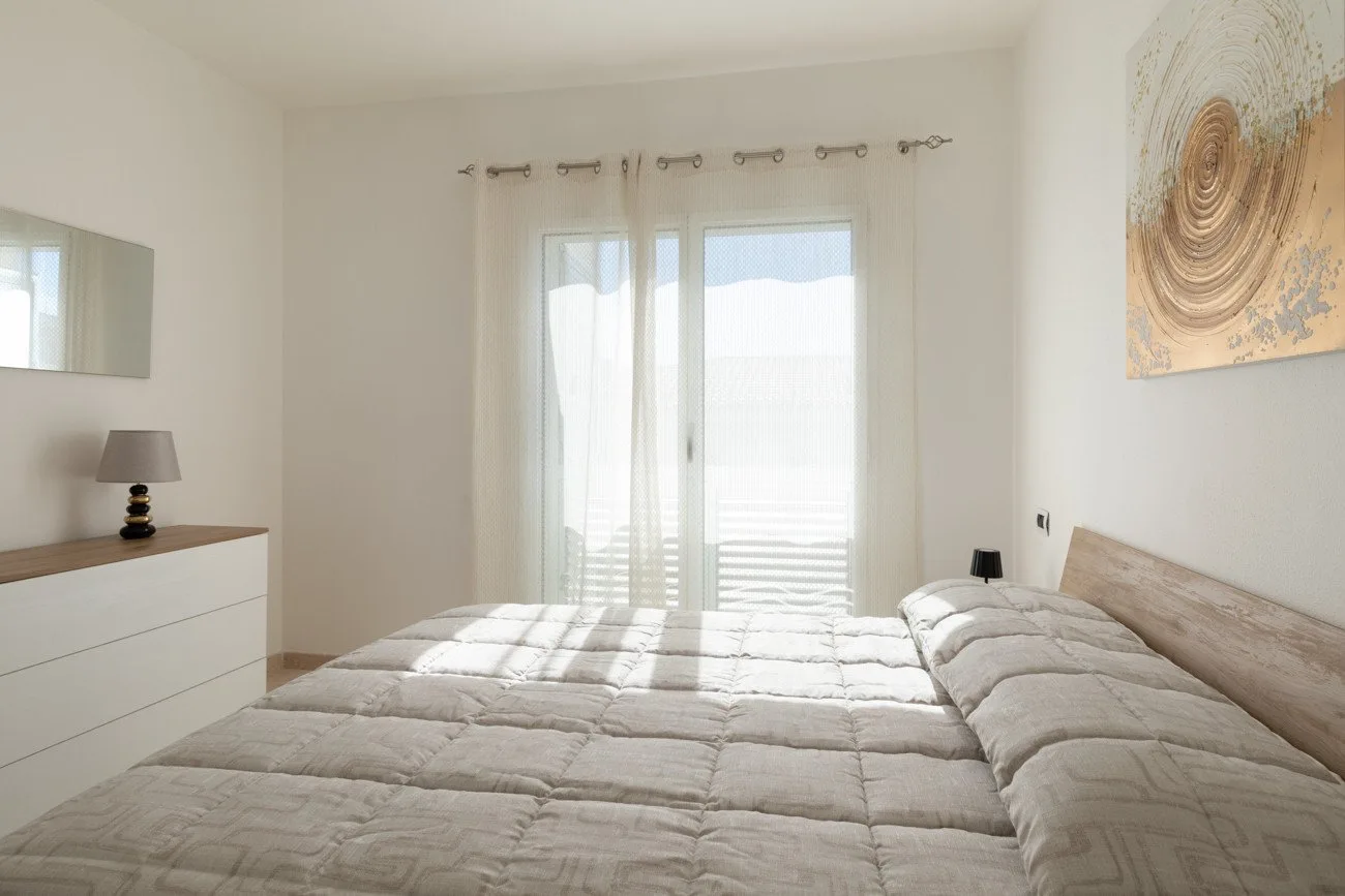 Un dormitorio luminoso con una cama grande junto a puertas de vidrio y cortinas translúcidas.