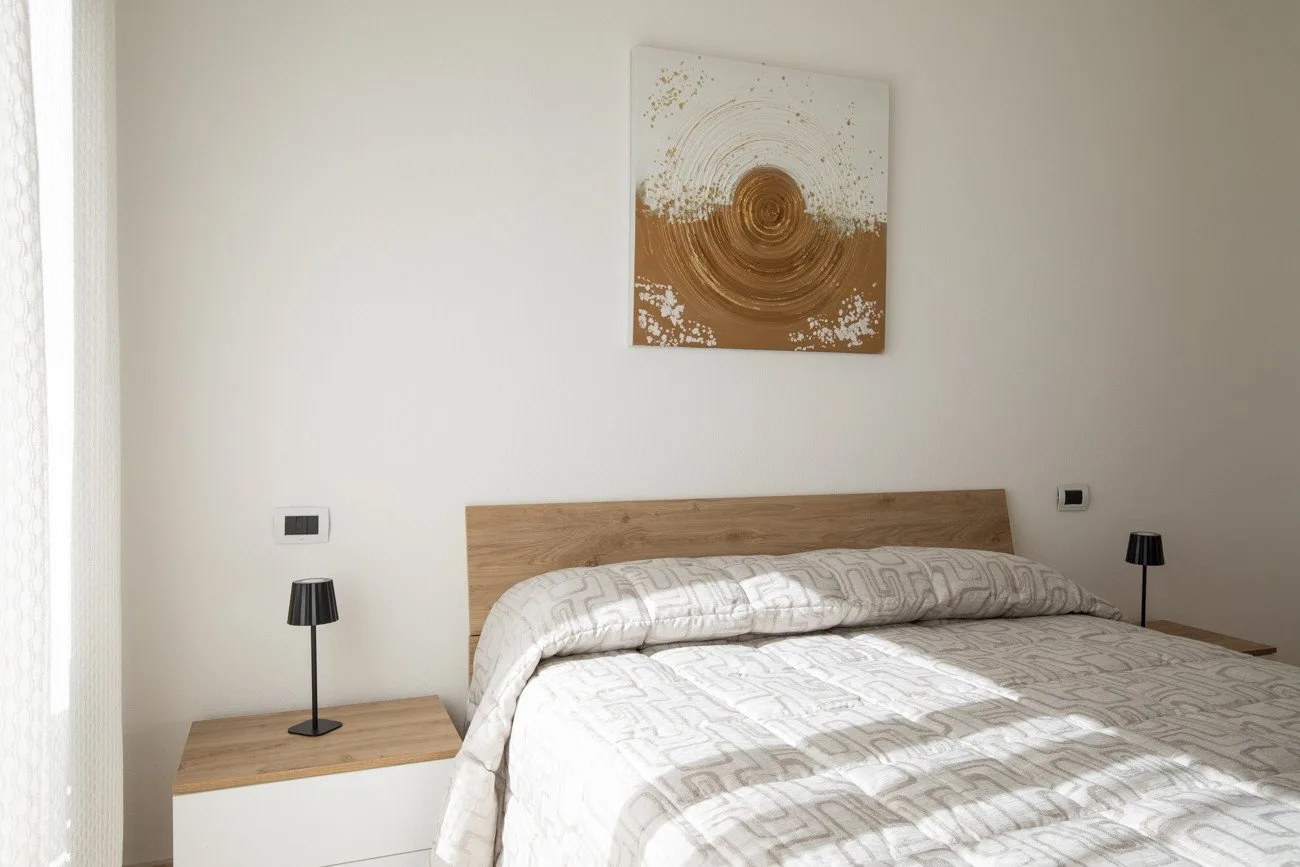 Un dormitorio con una cama bajo un cuadro abstracto y una ventana luminosa.