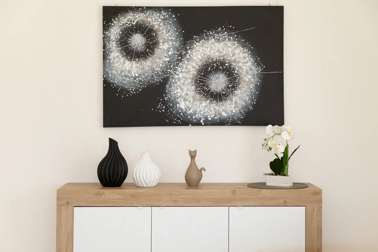 Una consola de madera clara con jarrones decorativos y flores está debajo de una pintura abstracta negra.