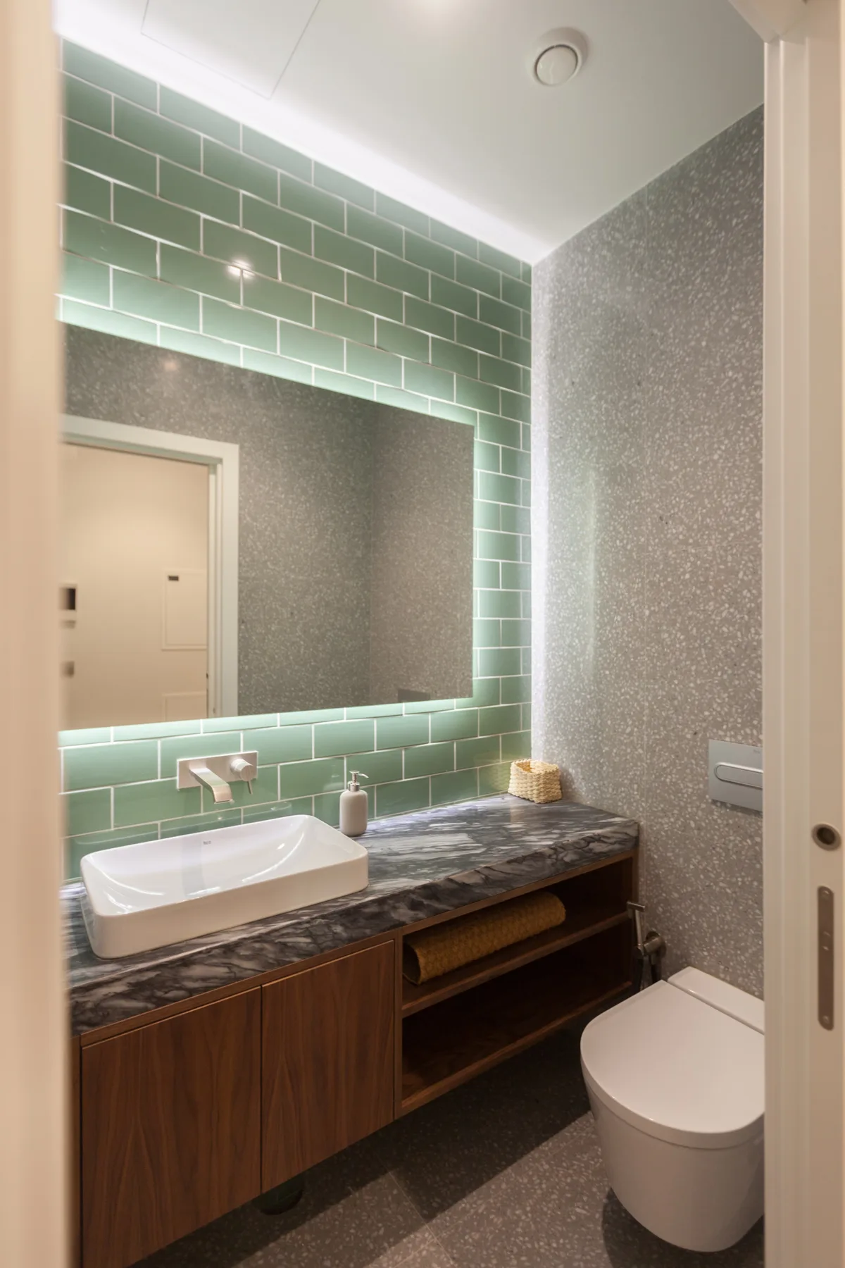 A bathroom with a green-tiled mirror wall, vanity, and toilet.