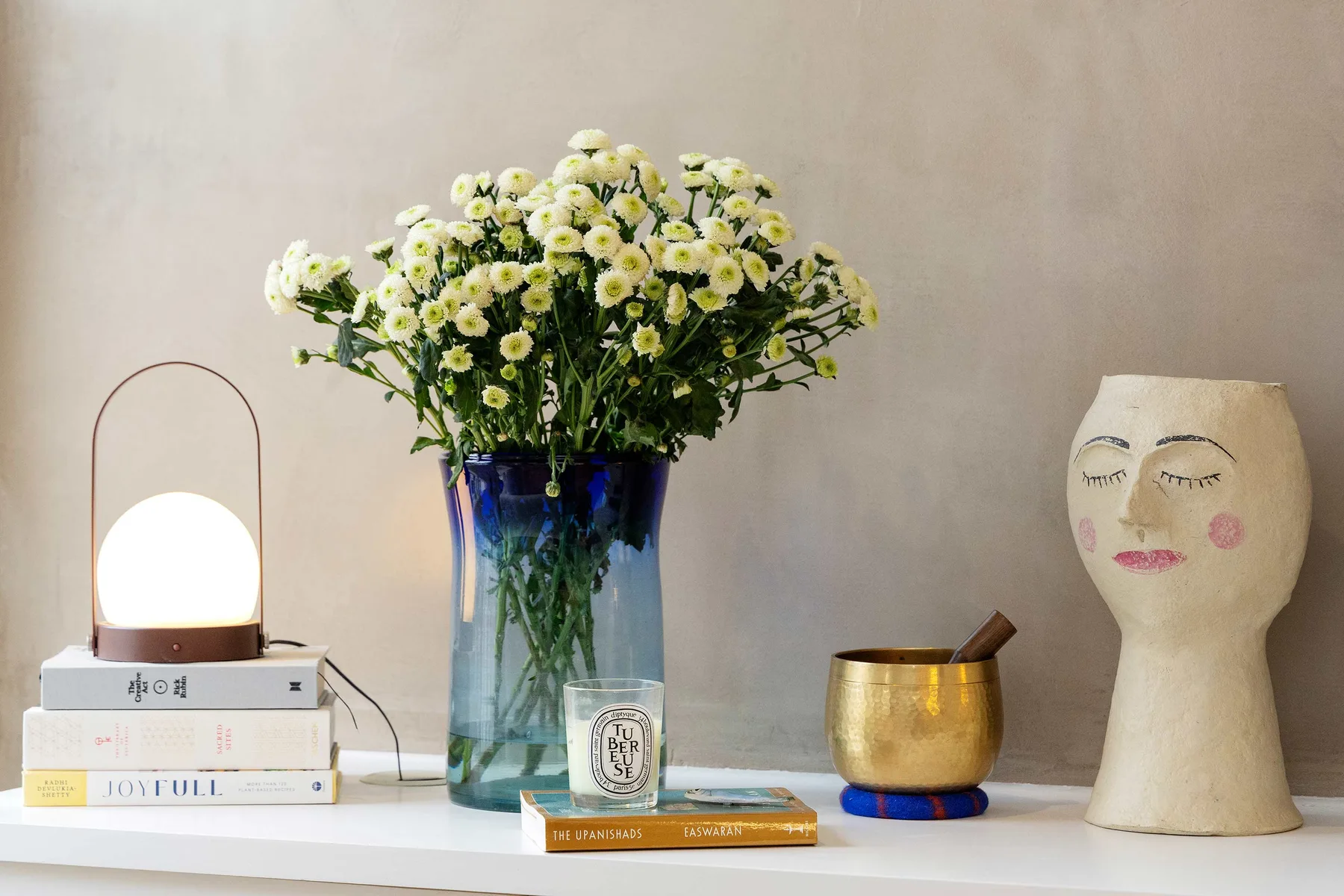 A tabletop still life features a vase of flowers, stacked books, and decorative objects.