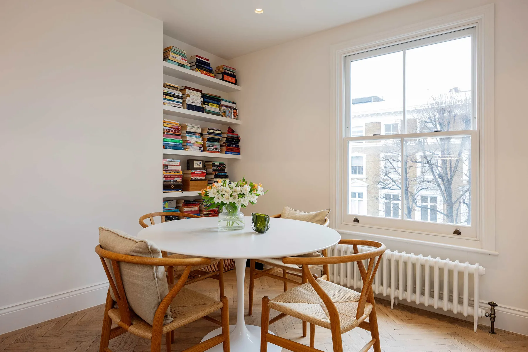 A bright dining nook has a round table, wooden seating, and shelves of books.