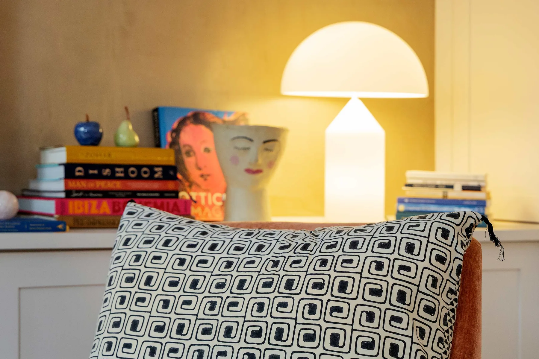 A warmly lit living room with a patterned cushion in front of books and artwork.
